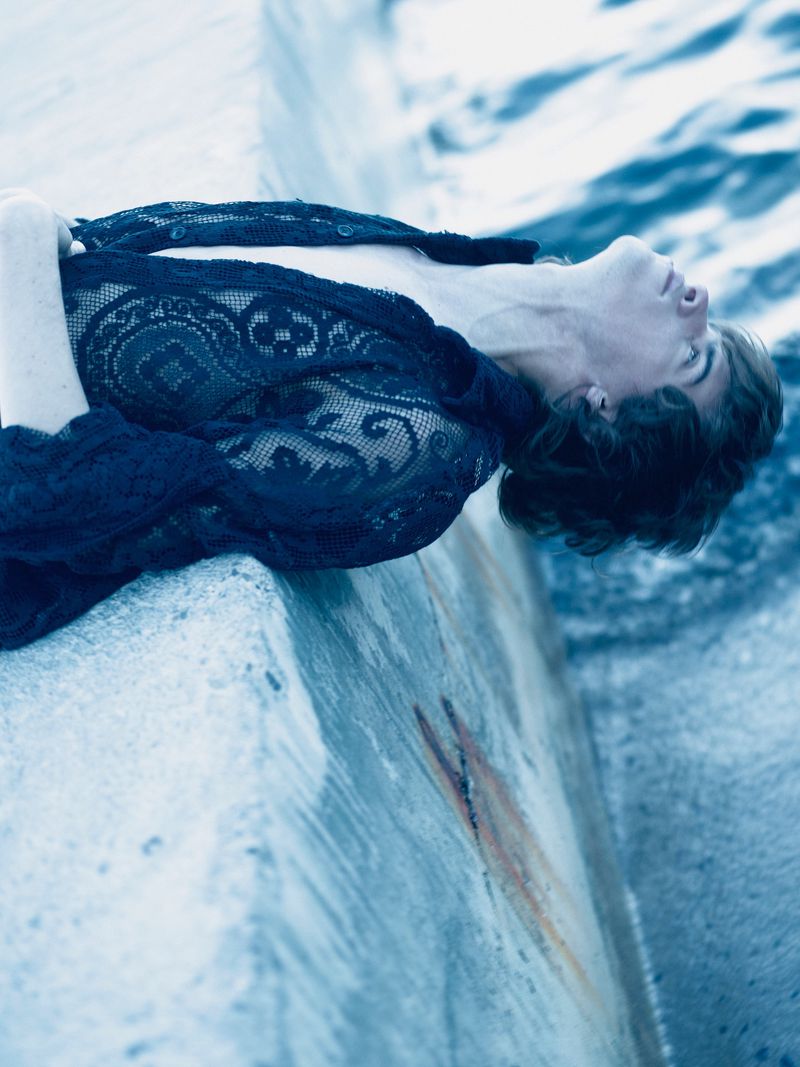 Adult rests on a concrete structure beside the ocean, wearing a lace top, in a medium shot, with cool blue tones and natural light.