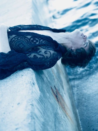 Adult rests on a concrete structure beside the ocean, wearing a lace top, in a medium shot, with cool blue tones and natural light.