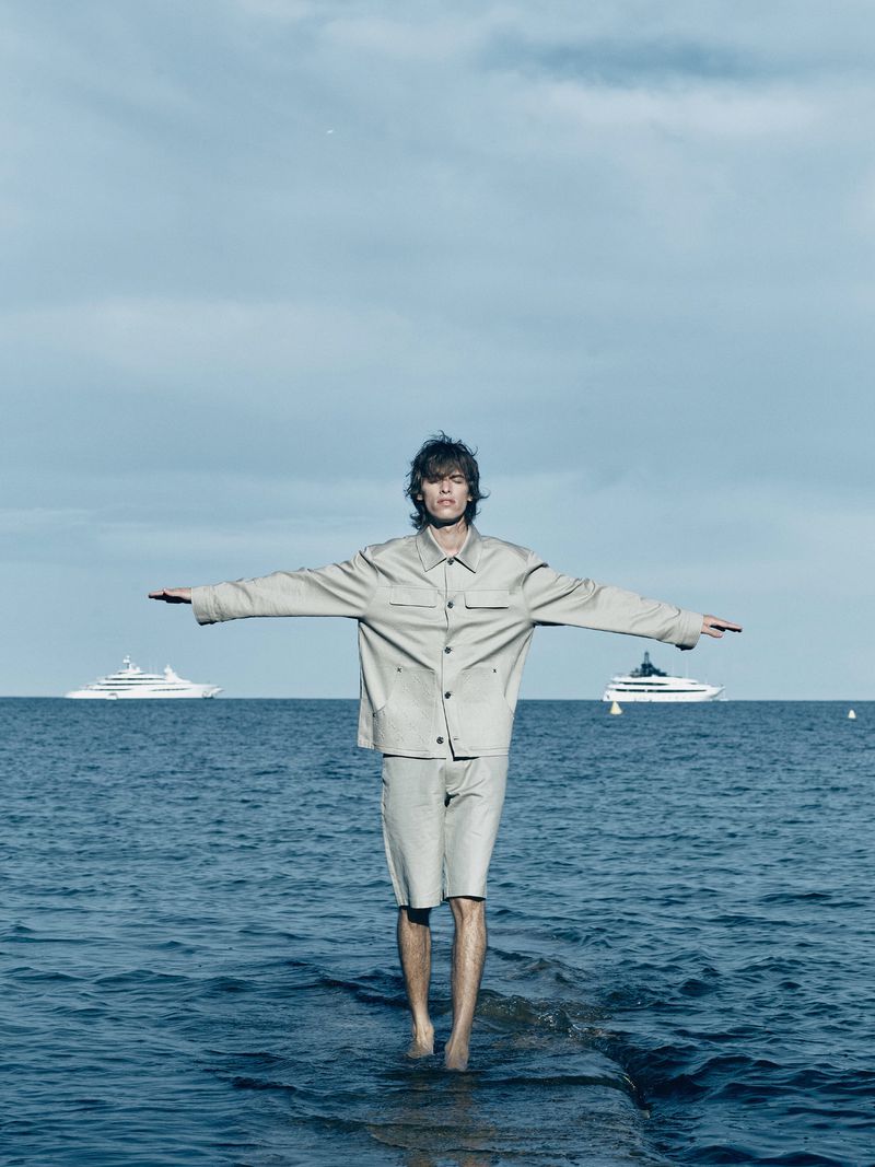 Man in a beige jacket and shorts stands barefoot on a rock formation in the ocean with arms outstretched, boats in the background.