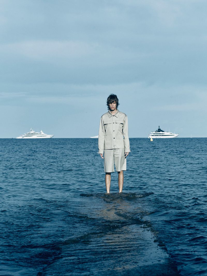 A person in a light-colored jacket and shorts stands waist-deep in ocean water on a rocky patch with yachts in the background under a blue sky.