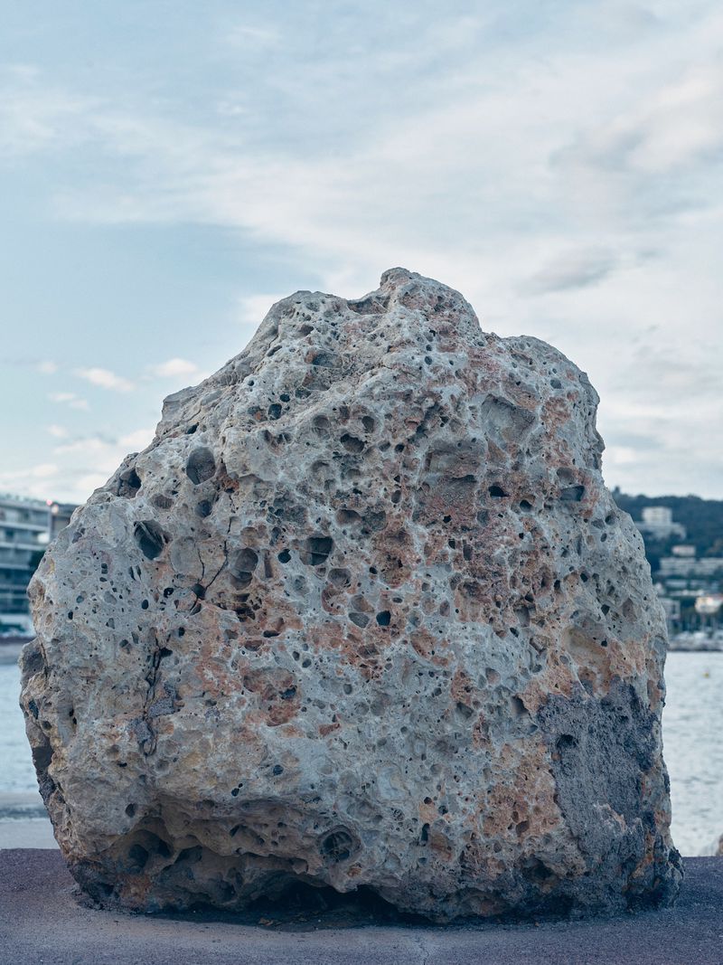 Large porous rock formation sits on a stone ledge, overlooking the ocean and coastline under a cloudy sky.
