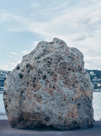 Large porous rock formation sits on a stone ledge, overlooking the ocean and coastline under a cloudy sky.