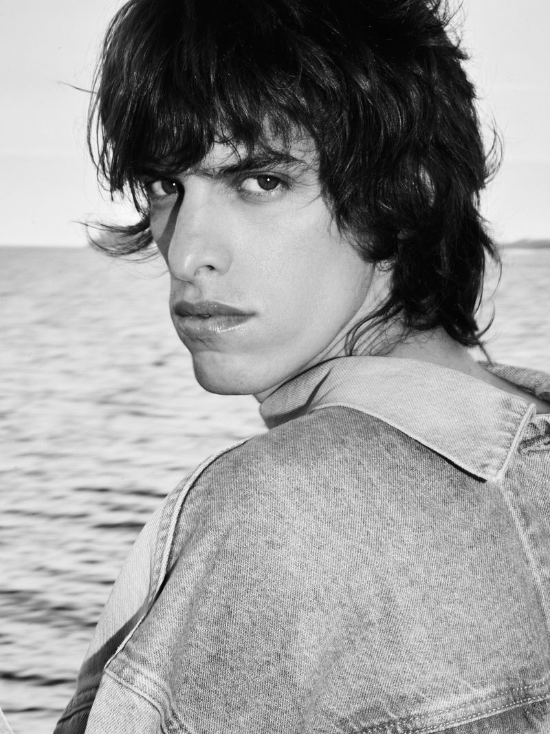 Black and white portrait of a young adult male with dark curly hair wearing a denim jacket in front of a seascape.