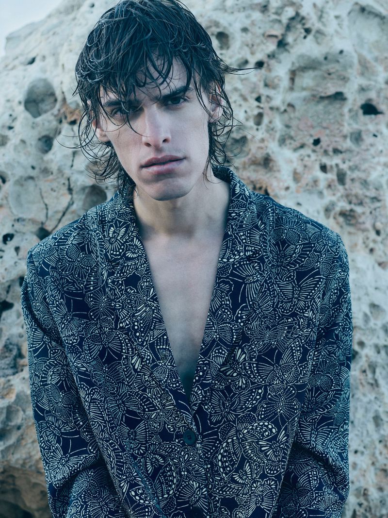Portrait of a male model with wet hair and a patterned blazer posing against a light rock formation in an outdoor setting.