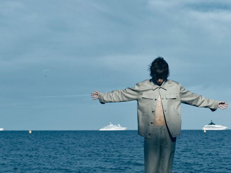 Adult stands on the beach with arms outstretched, wearing an open tan jacket and matching pants. Yachts sail in the background.