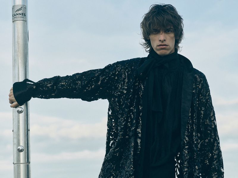Fashion shot of a male model in a black, floral-patterned coat leaning on a silver pole outdoors against a partly cloudy sky.