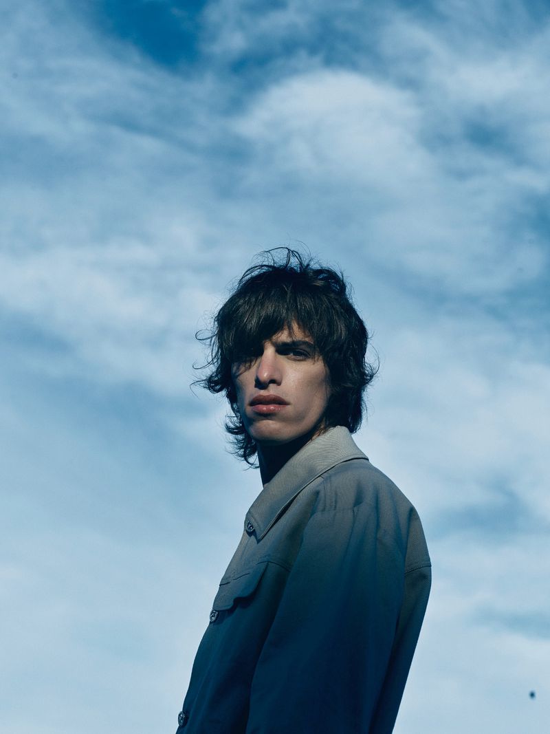 Low-angle portrait of a young adult male with dark hair and a grey jacket, set against a bright, cloudy blue sky outdoors.