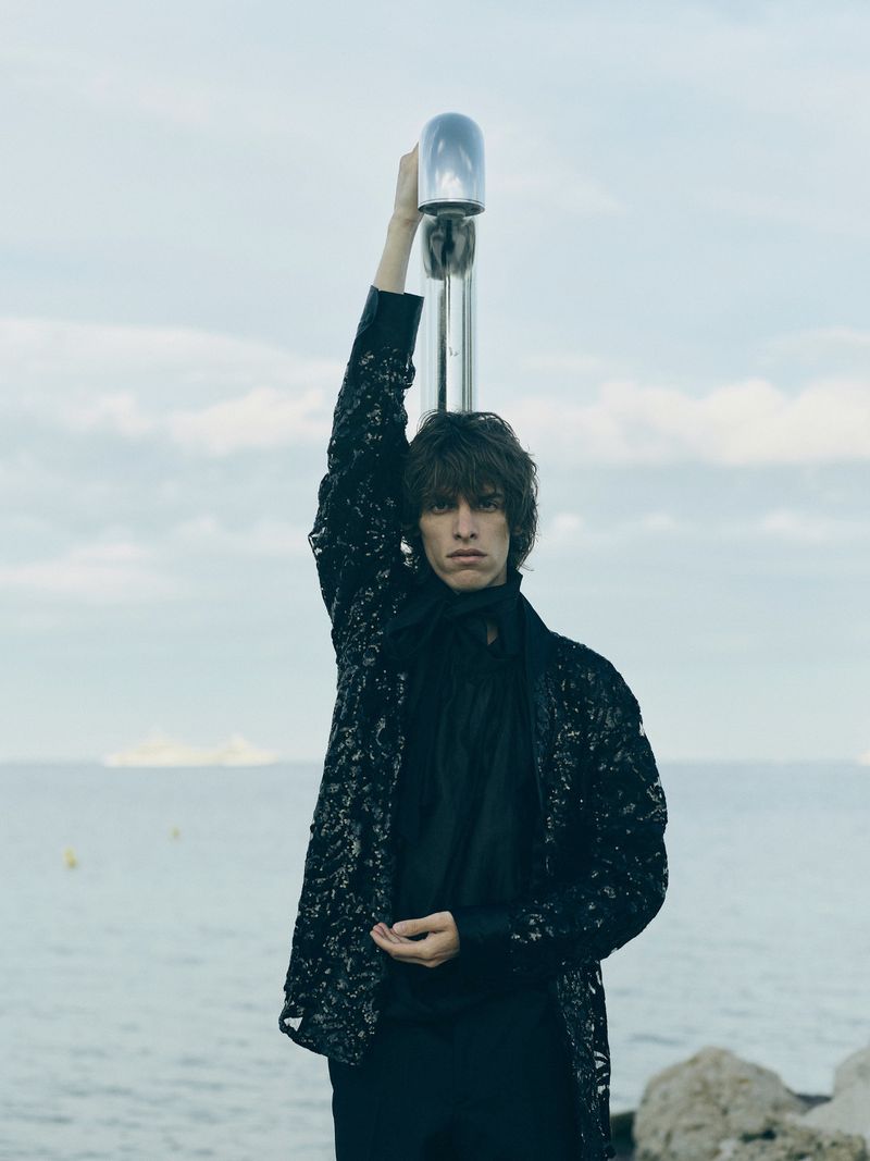 A stylish young adult man in a sequined jacket poses holding a metal structure near the ocean, creating a striking fashion moment.