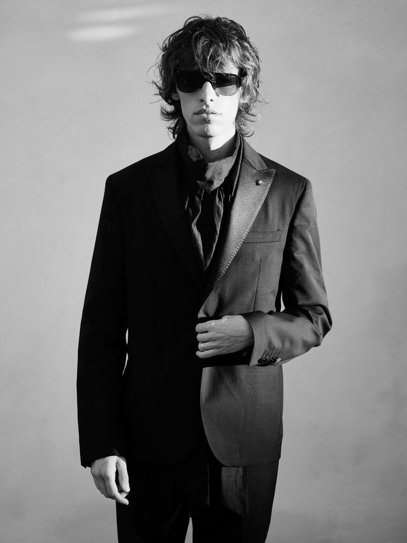 Black and white shot of a stylish adult man wearing a dark suit with a scarf and sunglasses in a bright, minimalist indoor setting.