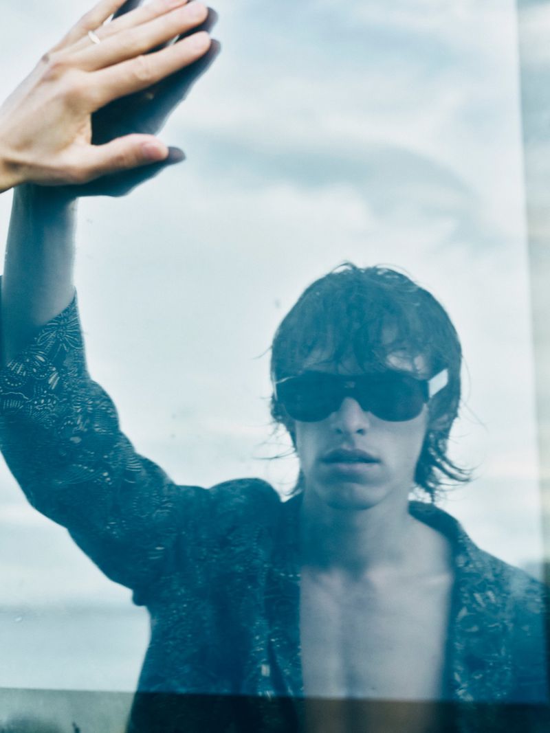 Man in sunglasses looking through glass panel with hand on it, cloudy sky, blue tones, fashion editorial, outdoor setting.