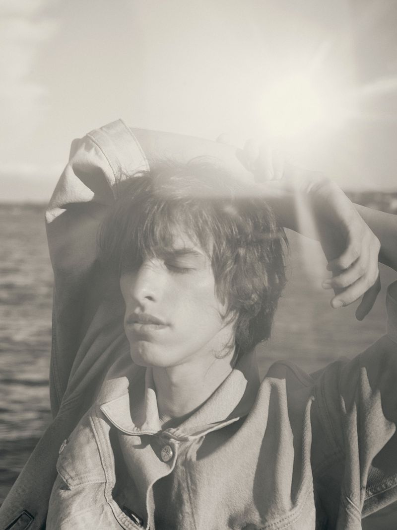 Portrait of a young adult male in denim jacket with closed eyes and hands behind his head, with a bright sunny seascape background.