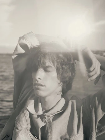 Portrait of a young adult male in denim jacket with closed eyes and hands behind his head, with a bright sunny seascape background.