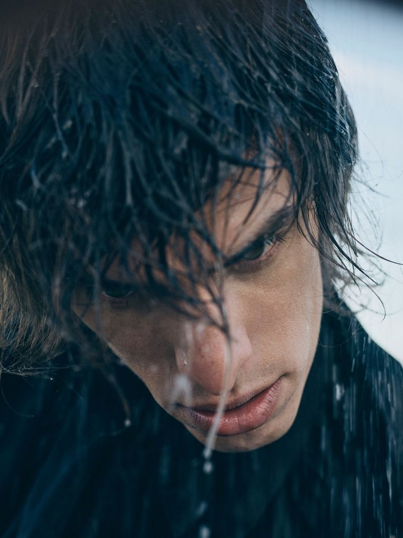 Close-up portrait of a young adult male with dark hair and light skin, wet from rain, gazing intensely towards the camera.