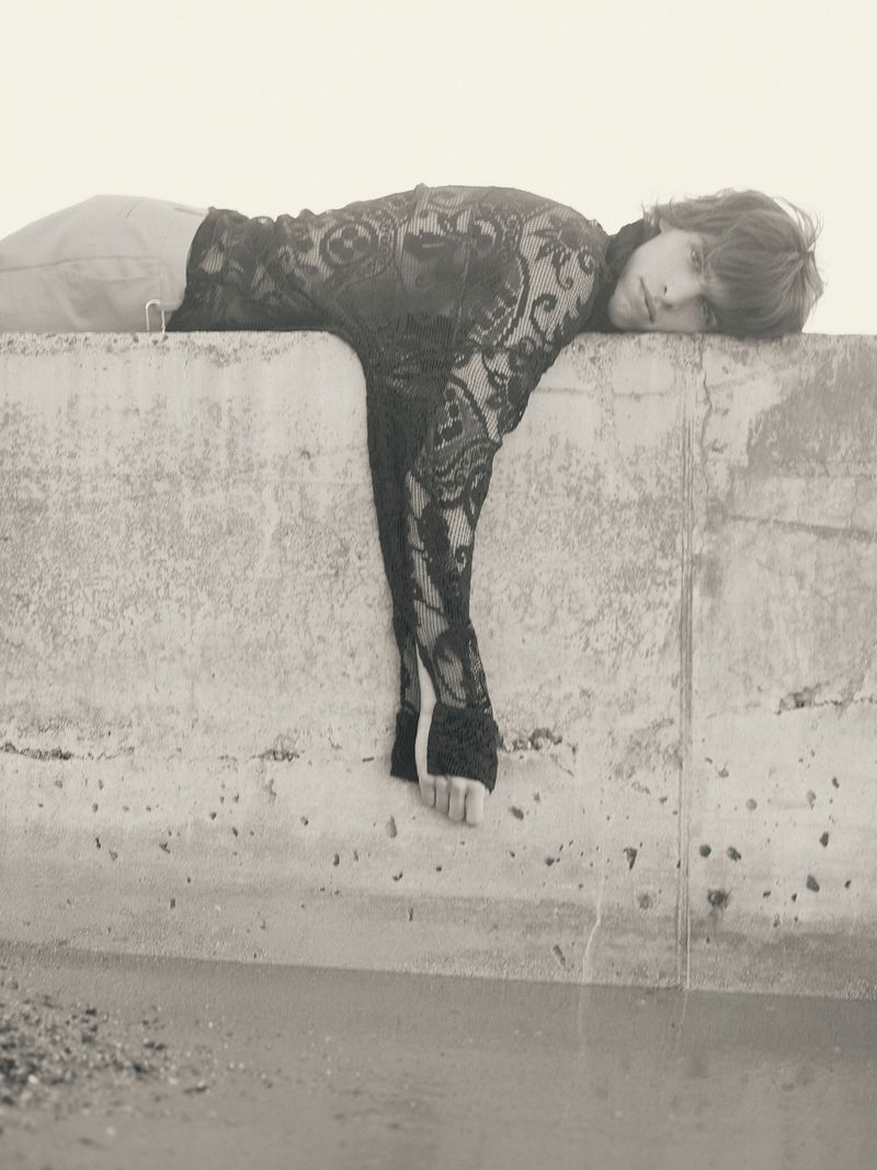 Monochrome portrait of a young adult with long hair wearing a lace top, lying on a concrete ledge in an outdoor setting near water.