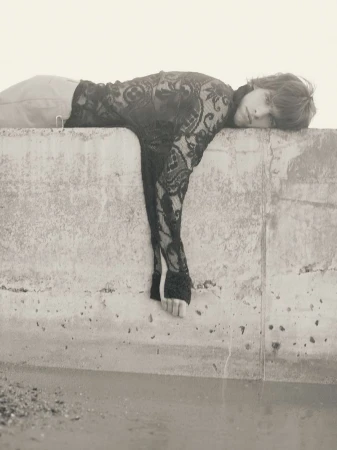 Monochrome portrait of a young adult with long hair wearing a lace top, lying on a concrete ledge in an outdoor setting near water.