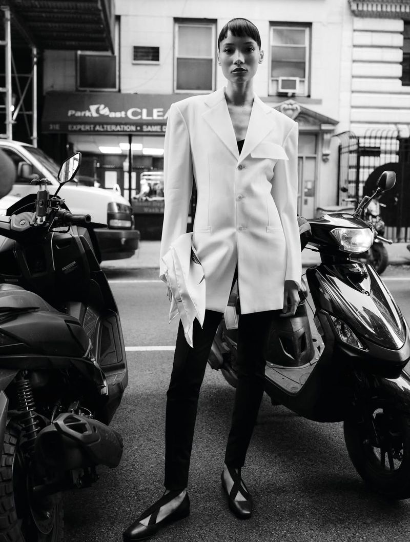 Monochrome portrait of a woman in a white blazer and black pants standing in front of scooters on a city street in natural lighting.