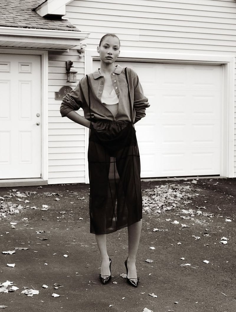 Adult female fashion model poses in a sheer skirt, button-down shirt and heels in front of white residential house, creating an editorial fashion shot.