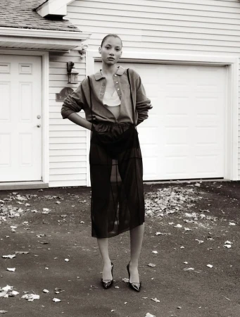 Adult female fashion model poses in a sheer skirt, button-down shirt and heels in front of white residential house, creating an editorial fashion shot.