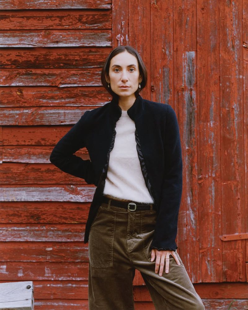 Adult woman poses in front of weathered, red wood wall wearing a black jacket, cream sweater, and olive-green corduroy pants.