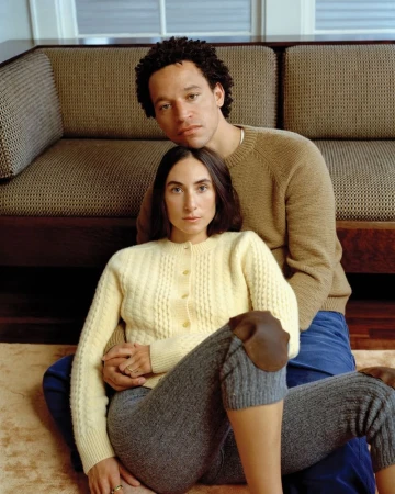 A couple is sitting indoors near a couch. The woman is wearing a yellow cardigan, and the man is wearing a tan sweater. The setting is warm.