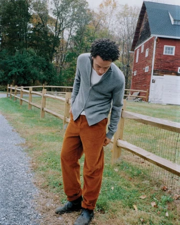 Adult male in casual attire poses by a wooden fence, with a red barn and trees in the background, captured in natural light.