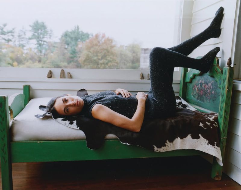 Woman in glittering top and tights lies on a green bed with a cowhide, posing in front of a bright window in an indoor setting.