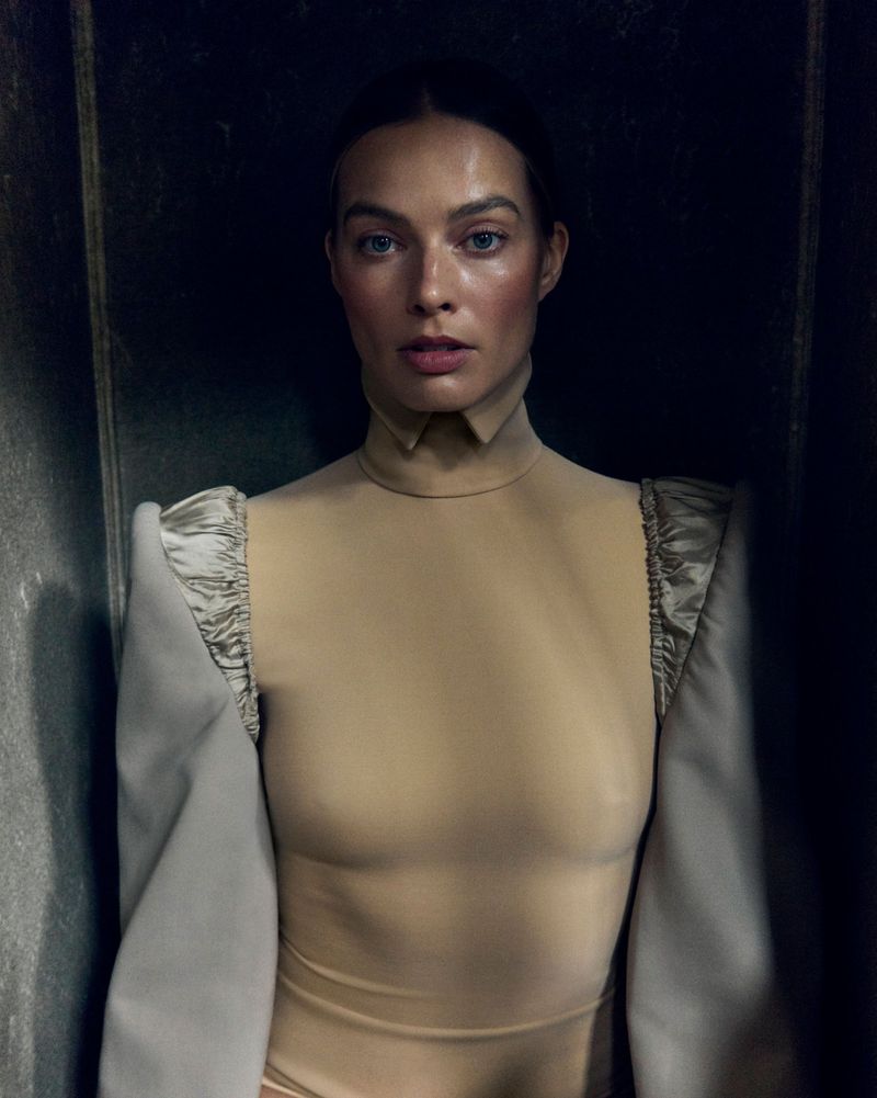 A portrait of an adult female model with fair skin, neutral expression, and light brown hair, wearing a beige bodysuit with structured shoulders.