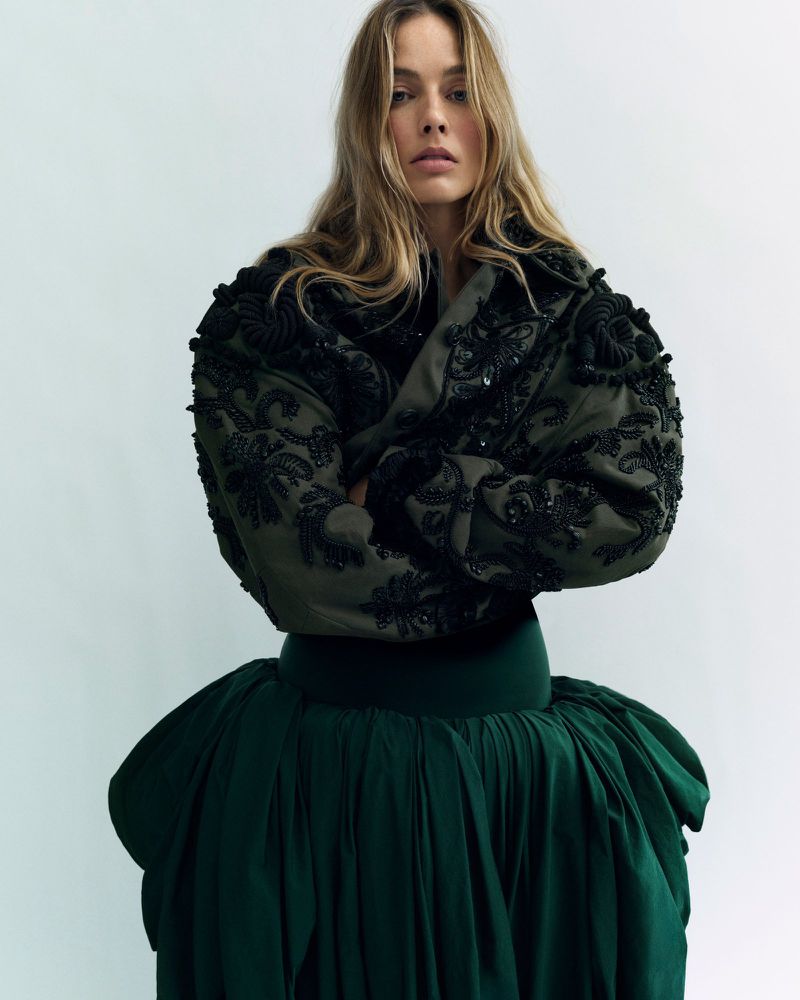 Portrait of a blonde woman in a green jacket and dark green skirt against a plain background in a fashion editorial style.