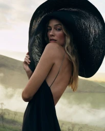 Margot Robbie wearing a large black wide-brim hat and dark dress outdoors in a landscape setting for British Vogue.