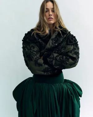 Margot Robbie in an embroidered dark jacket and voluminous emerald green skirt against a pale studio backdrop.