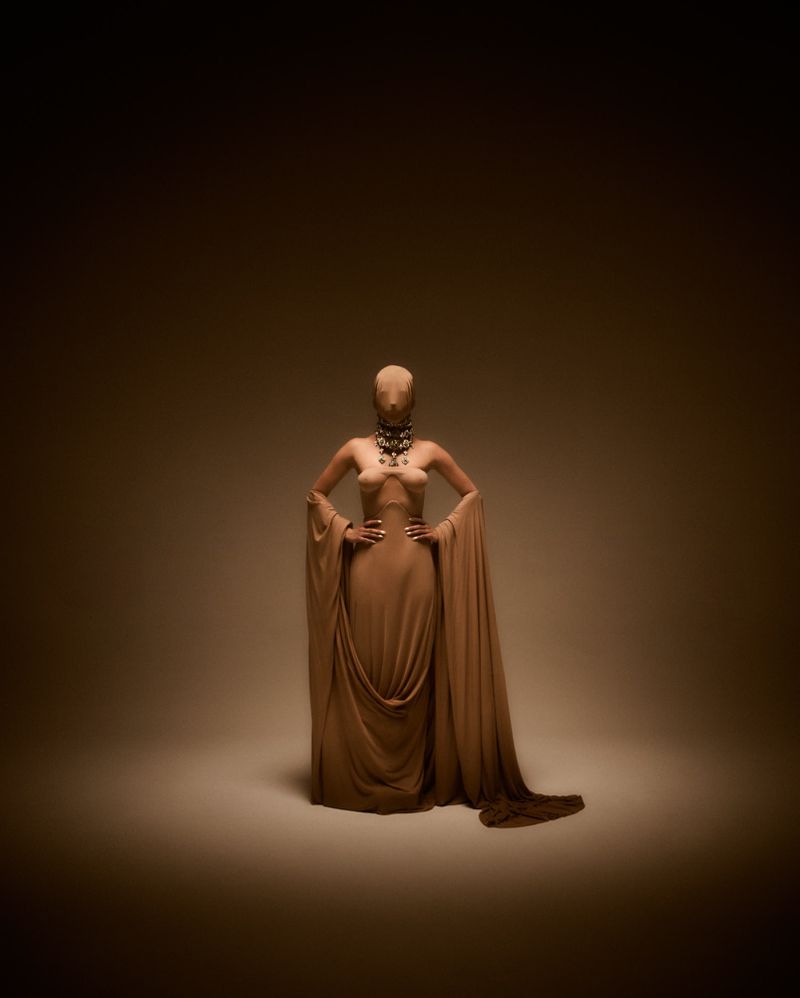 Woman in a monochrome sand-colored dress with a hood and long draped sleeves, standing in a spotlight against a brown background.