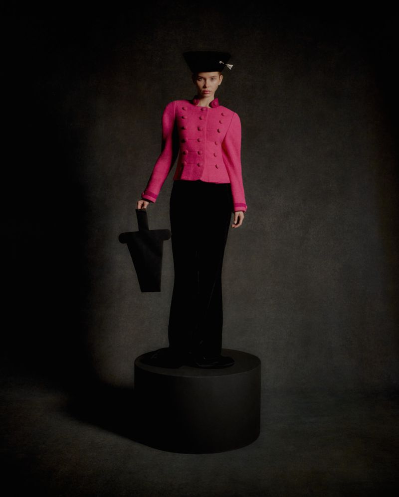A stylish female model poses on a platform in a pink jacket, black pants, and hat, holding a black geometric bag in a studio setting.