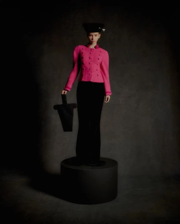 A stylish female model poses on a platform in a pink jacket, black pants, and hat, holding a black geometric bag in a studio setting.