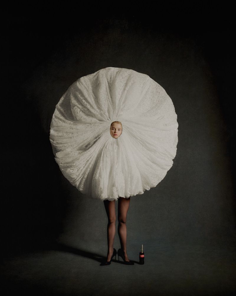 Artistic fashion portrait of a woman in a white tulle garment, black tights and heels standing next to a Coca-Cola bottle against a grey background.