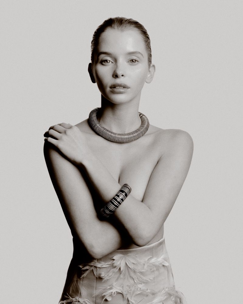 Woman posing with crossed arms wearing a chunky ribbed collar necklace, patterned cuff bracelet, and feather skirt on a studio backdrop.