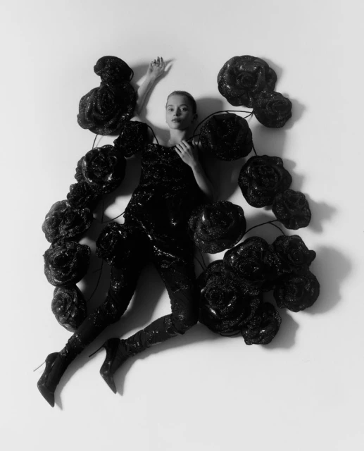 Monochrome portrait of a woman in a glittery outfit lying among a scattered display of black rose-like forms against a bright backdrop.