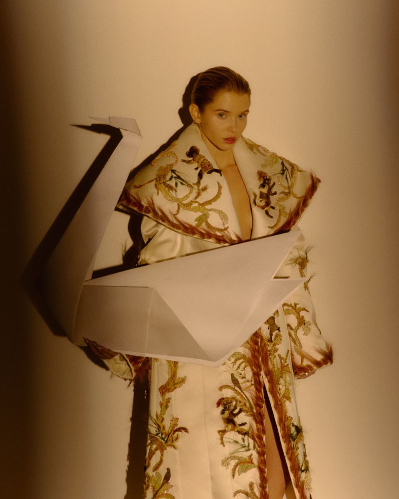 Woman in embroidered white coat holding large paper crane in photo studio with soft lighting, fashion editorial.