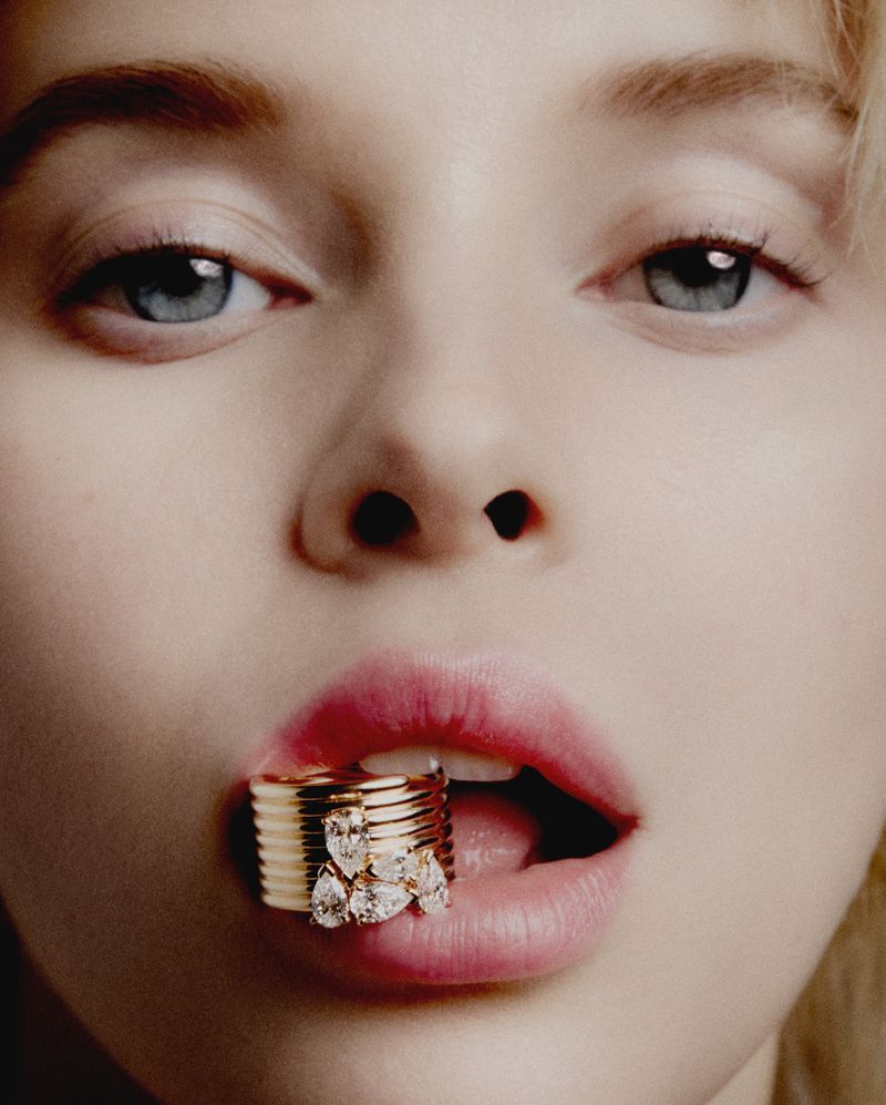Close-up portrait of a blond woman with a gold ring in her mouth, showcasing beauty and jewelry in a studio setting, emphasizing facial features.