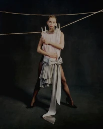Model posing behind a pleated pastel garment pinned with wooden clothespins to a rope against a dark studio background.