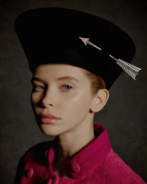 Close-up portrait of a woman wearing a black hat with a silver arrow pin and a pink textured jacket against a dark textured background.