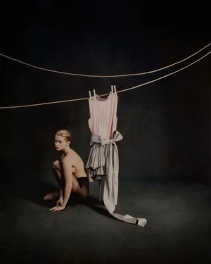 Model crouched on dark floor beside a dress hanging from ropes for Vogue Portugal editorial