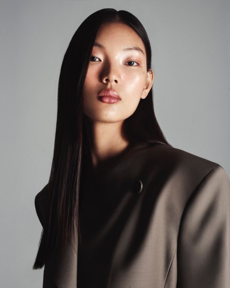 Close-up portrait of an Asian woman wearing a brown blazer against a soft grey backdrop in a studio fashion shot.