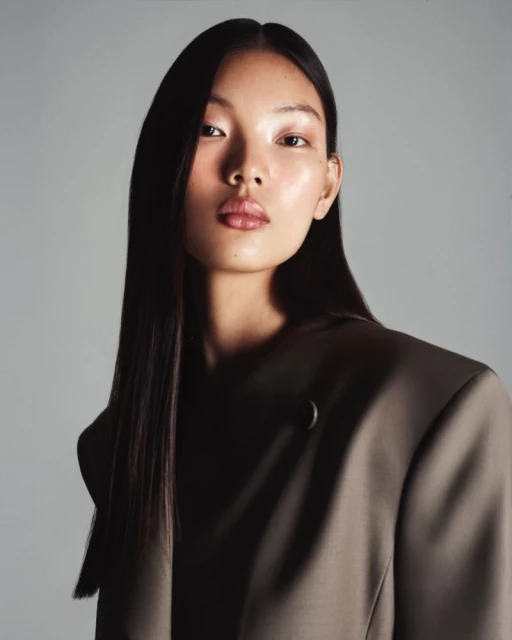 Close-up portrait of an Asian woman wearing a brown blazer against a soft grey backdrop in a studio fashion shot.