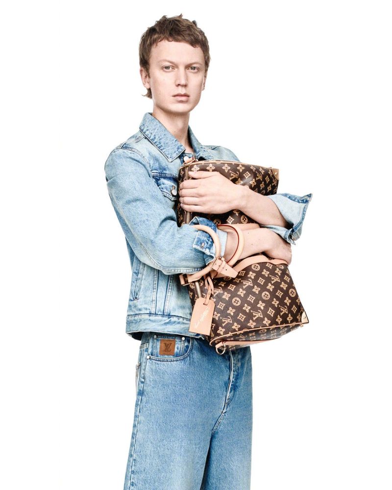 A light-skinned person with short hair wearing a denim jacket and light denim jeans holding two brown monogrammed handbags in a studio setting.