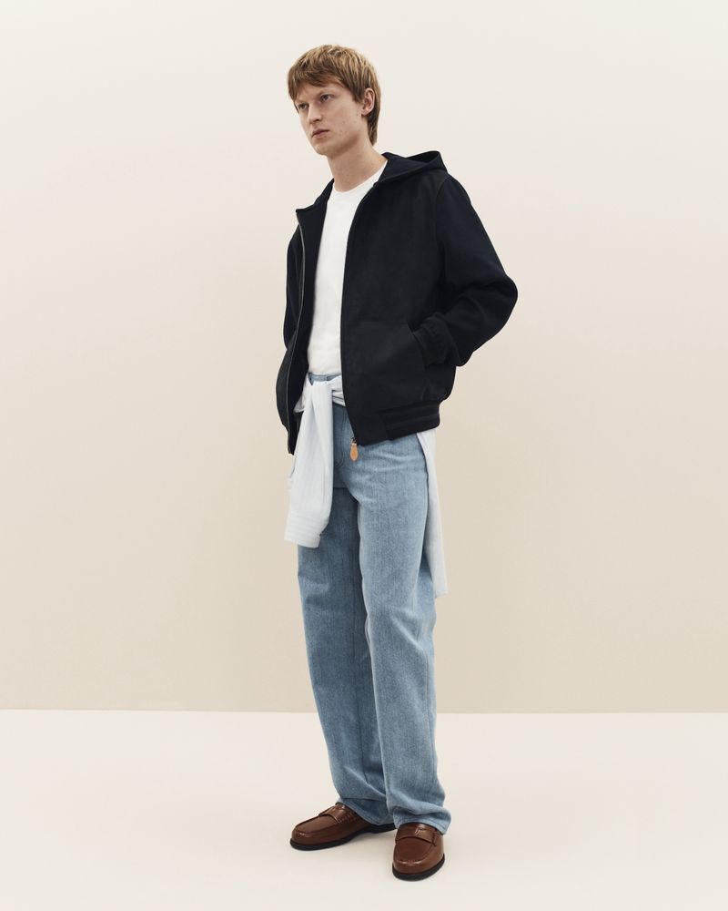A stylish ginger-haired man models apparel in a studio, wearing a dark jacket, t-shirt, jeans, and loafers against a neutral backdrop.