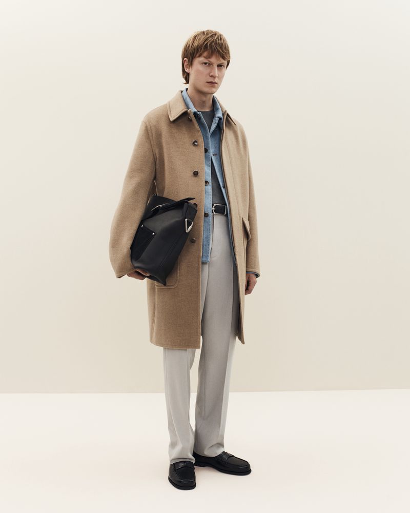 Man in tan coat with black bag stands in a studio setting. He is wearing a blue shirt, grey pants, and black loafers.