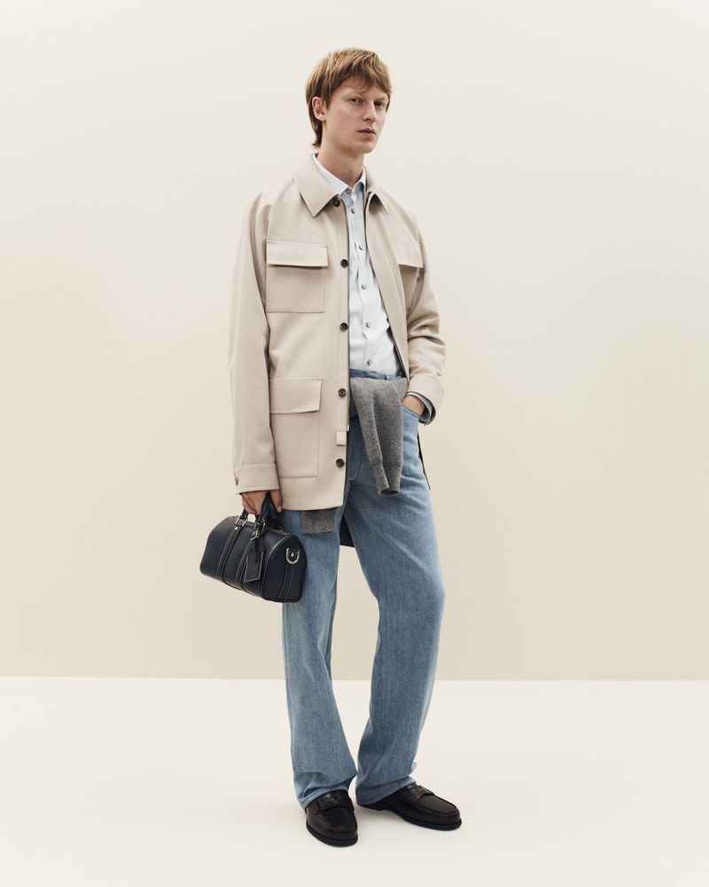 Male model wears a beige overshirt, blue jeans, and black loafers, holding a duffel bag in a fashion studio shot.