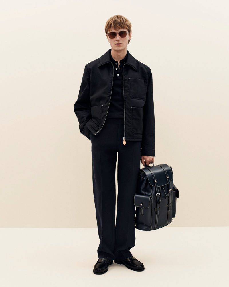 A fair-skinned adult male stands against a cream background wearing a navy jacket, matching trousers, and loafers, holding a navy backpack.