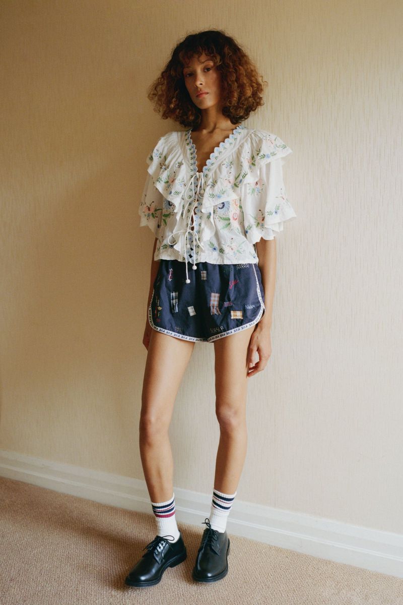 Full shot of a woman in a white floral blouse and dark shorts, posed indoors, with black shoes and ankle socks with colored stripes.