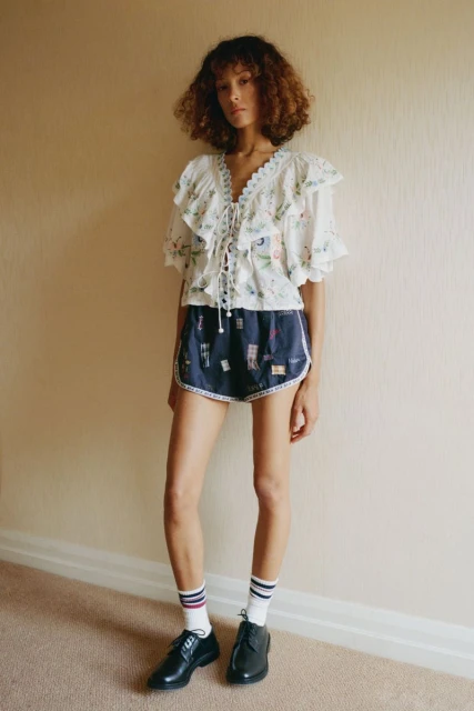 Full shot of a woman in a white floral blouse and dark shorts, posed indoors, with black shoes and ankle socks with colored stripes.